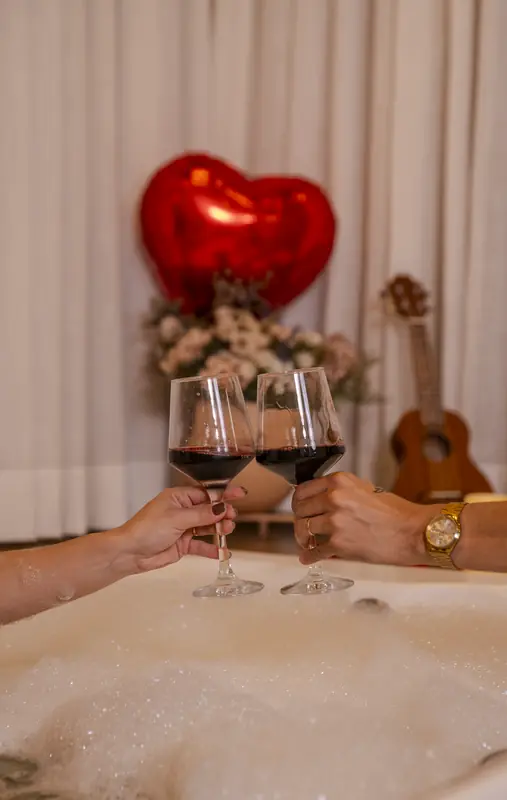 Casal brindando com vinho dentro da banheira de hidromassagem, com um balão de coração ao fundo