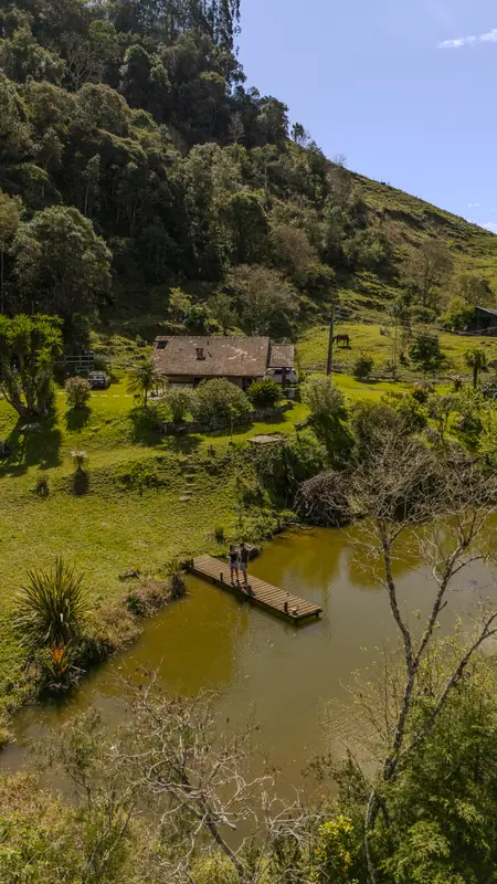 O lago com o deck e a casa no alto, cercados de mata